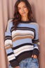 A woman wearing a knit crewneck sweater with horizontal stripes in light blue, cream, tan, and dark brown, paired with blue jeans, posed against a draped dusty pink backdrop.