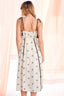 Woman wearing a white dress with black floral patterns and thin straps against a light pink curtain background.