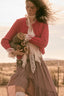 A woman outdoors wearing a red cable-knit cardigan, a white lace blouse, and a brown-and-white plaid skirt, holding a small stuffed animal. The background is a softly blurred warm-toned landscape.