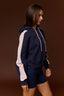 A woman wearing a navy blue colorblock hoodie with light pink sleeve stripes and matching navy shorts, posed against a solid dark brown studio background.