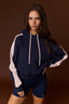 A woman modeling a navy blue hoodie with light pink sleeves and matching navy athletic shorts against a solid dark brown studio background.