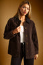 A woman wearing a dark brown corduroy jacket over a white tank top and black pants, posed against a warm brown studio background.