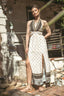 A woman models a sleeveless bohemian-style maxi dress with a high slit, featuring a cream and brown geometric pattern. She is standing outdoors on a brick patio in front of a rustic stone wall.