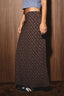 A person wearing a long dark plaid skirt with brown and red lines, paired with a blue crop top and black sneakers, standing against a dark wood-paneled wall.