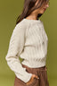 A woman wearing a cream cable-knit sweater and brown pants, shown in profile against a solid olive green background.