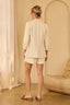 Woman wearing a beige outfit standing in a room with a beige wall and curtain.