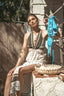 A woman sits outdoors on a stone ledge wearing a patterned halter maxi dress, holding a turquoise bikini top on a clothesline. A woven basket sits on the stone beside her, with a wooden fence and outdoor setting in the background.