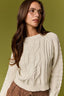 A woman wearing a cream cable-knit sweater and brown pants, standing against a solid olive green background.