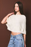 A woman modeling a cream-colored, cropped cable-knit sweater with wide sleeves, paired with light-wash blue jeans, set against a solid dark brown studio background.
