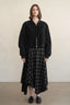 A model stands against a plain beige wall wearing a black cropped zip-front jacket with voluminous sleeves, paired with a black-and-gray plaid midi skirt and black slip-on shoes.