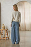 Woman wearing a beige sweater and blue jeans standing in a room with a wooden stool and ceramic vase.