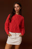 A woman wearing a red cable-knit cropped sweater and a white mini skirt, standing against a solid dark brown studio background.