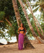 Woman in a black top and purple sarong standing under palm trees by the beach.