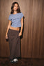 A woman modeling a light blue crop top with 'good vibes' printed in black script, paired with a dark brown and red plaid wrap skirt and black sneakers, standing against a brown wood-paneled wall.