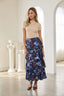 A woman stands in a bright white room with arched windows and columns, wearing a beige short-sleeve top and a navy blue tiered maxi skirt with a colorful floral and abstract print.