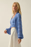 Woman wearing a blue knitted cardigan over a crop top with white pants on a plain background