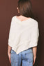 A woman shown from the back wearing a cream-colored, cable-knit wrap poncho over a V-neck top, paired with blue jeans, against a solid dark brown background.