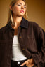 A woman wearing a dark brown corduroy jacket with a contrasting dark navy collar, layered over a white tank top and black jeans, set against a warm brown studio background.