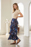 A woman modeling a navy blue tiered maxi skirt with a colorful floral and abstract print, paired with a beige short-sleeve top and brown flats, standing in a bright, modern white room with arched windows.