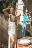 A woman stands outdoors wearing a sleeveless bohemian-style two-piece set with a patterned cream and black skirt and a matching halter top. A bright blue bikini hangs on a clothesline next to her, with a woven basket and rustic stone wall in the background.