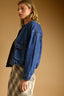 A woman wearing a cropped blue denim jacket over a black top and a beige plaid skirt, photographed against a warm tan studio background.