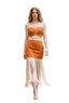 A woman models a two-piece orange and cream fringe outfit consisting of a crop top and a high-waisted skirt, set against a blurred background of a brick archway and stone walkway.