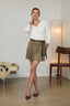 A woman models a white long-sleeve knit top and an olive green suede mini skirt with a matching tie belt, styled with brown pointed-toe flats in a modern indoor setting.
