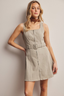Woman wearing a beige sleeveless dress with a belt against a plain background