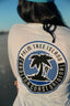 Person wearing a white shirt with a palm tree design and text on the back, standing on a beach.