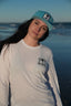 Woman wearing a white long-sleeve shirt and blue cap with a logo, standing on a beach.